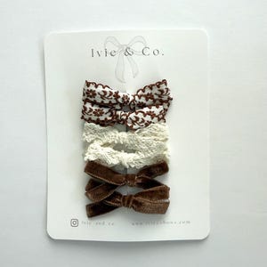 May include: A set of four hair bows attached to a white display card labelled Ivie & Co. The top two bows are white with a brown floral pattern and scalloped edges. The third is a cream lace bow, and the bottom two are solid brown velvet bows.