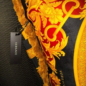 May include: A decorative pillow with a black background and a red and gold baroque design. The pillow has gold fringe trim and a black tag that reads "VERSACE".