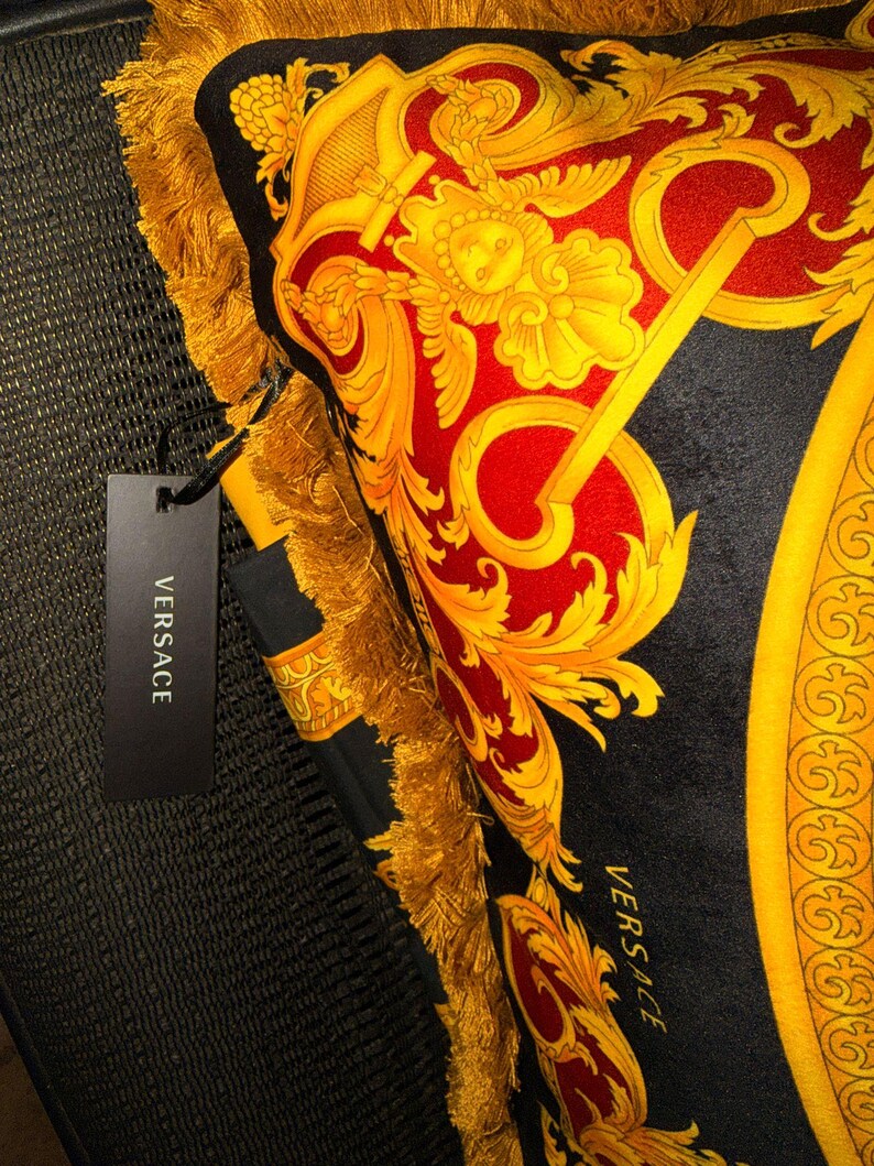 May include: A decorative pillow with a black background and a red and gold baroque design. The pillow has gold fringe trim and a black tag that reads "VERSACE".