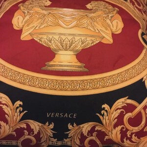 May include: A silk scarf with a bold design featuring a central gold-colored urn flanked by two lion figures. The urn is set against a red background, framed by a gold border and ornate black and gold flourishes. The word "VERSACE" is printed in white.