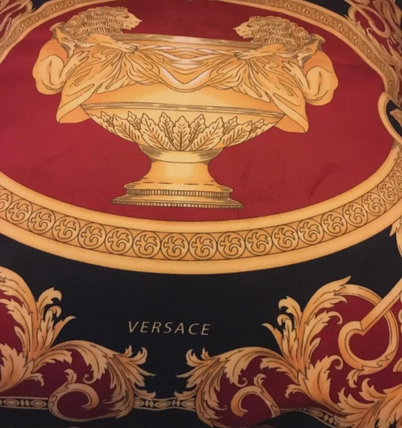May include: A silk scarf with a bold design featuring a central gold-colored urn flanked by two lion figures. The urn is set against a red background, framed by a gold border and ornate black and gold flourishes. The word "VERSACE" is printed in white.