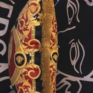May include: Decorative pillow with a black, red, and gold baroque pattern. The pillow features a gold fringe trim along one edge. The design includes ornate swirls, medallions, and a central panel with a repeating pattern.