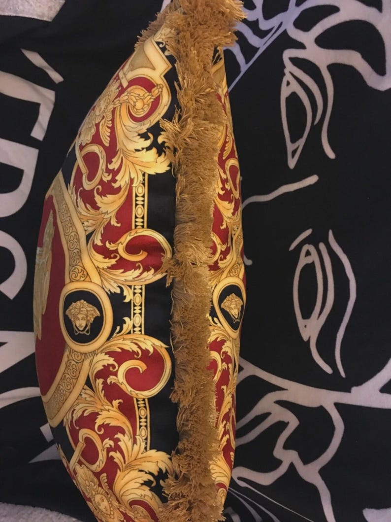 May include: Decorative pillow with a black, red, and gold baroque pattern. The pillow features a gold fringe trim along one edge. The design includes ornate swirls, medallions, and a central panel with a repeating pattern.