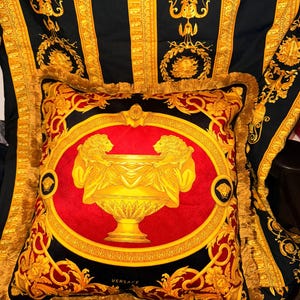 May include: A decorative pillow with a red center featuring a gold urn design flanked by two figures. The pillow is framed by a gold border and fringe, set against a black and gold patterned background.