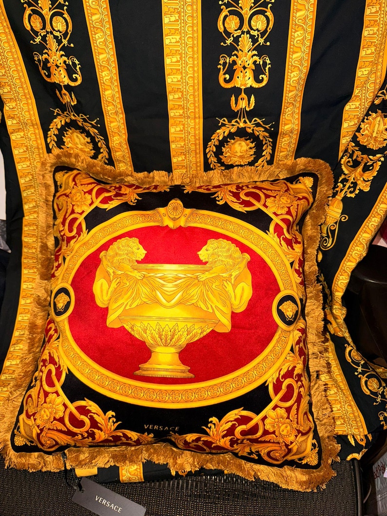 May include: A decorative pillow with a red center featuring a gold urn design flanked by two figures. The pillow is framed by a gold border and fringe, set against a black and gold patterned background.