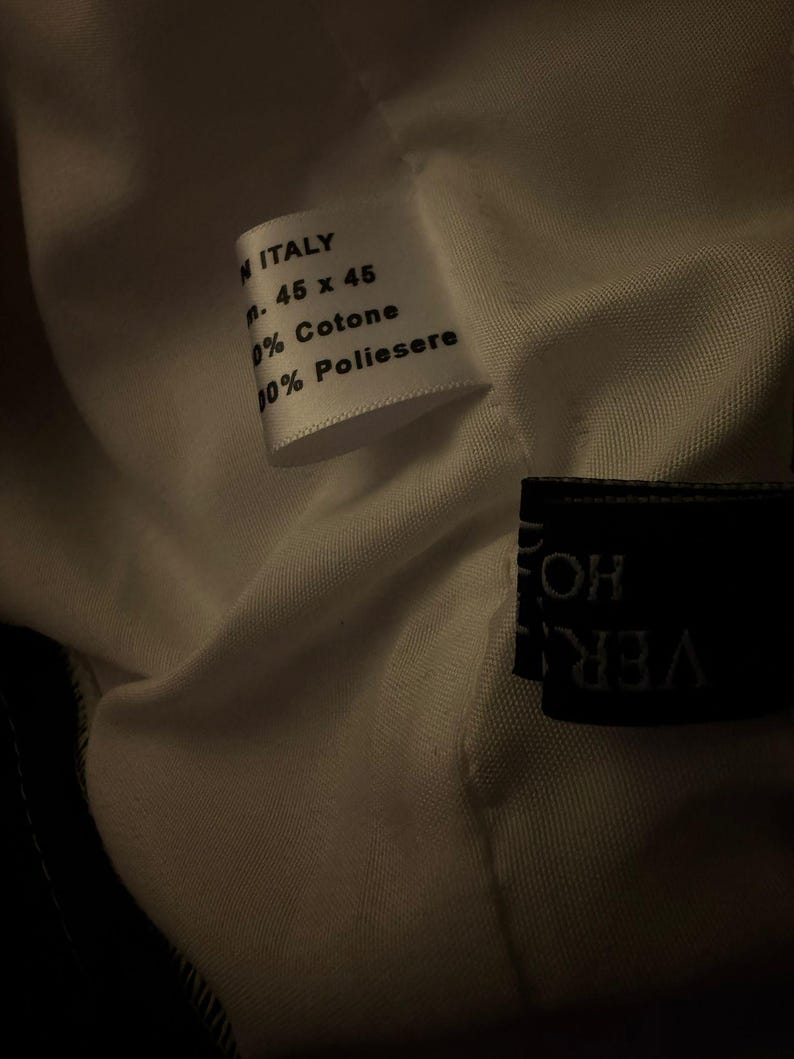 May include: Close-up of a white fabric tag with text. The tag reads "MADE IN ITALY", followed by measurements "cm. 45 x 45", and fabric composition details: "100% Cotone" and "100% Poliestere".