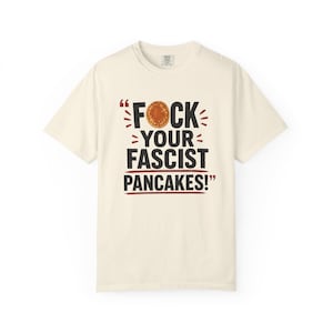 Puede incluir: Una camiseta de manga corta de color crema con el texto negro "FOCK YOUR FASCIST PANCAKES!", donde un panqueque dorado sustituye la letra O de la primera palabra, acentuado con comillas y líneas horizontales de color rojo.