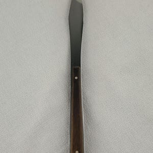May include: A stainless steel butter knife with a dark brown wooden handle. The knife has a pointed tip and two small white rivets on the handle. The knife is designed for spreading butter or other soft spreads.