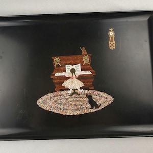 May include: A rectangular black tray with a decorative scene. The scene features a piano, a girl, a cat, and a clock. The piano is brown with gold accents. The girl is wearing a pink dress. The cat is black.