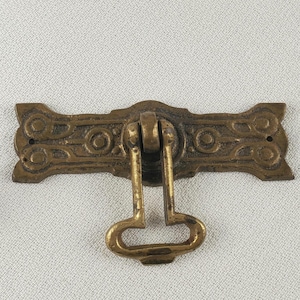 May include: A vintage-style brass drawer pull with an ornate, rectangular backplate and a drop handle. The backplate features a repeating pattern of circles and curves. The handle is a simple, open-ended design. The hardware is likely for a cabinet or drawer.