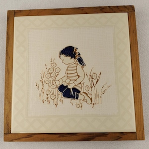 Può includere: Piastrella quadrata in ceramica con cornice in legno. La piastrella presenta un'illustrazione marrone e blu navy di un bambino che raccoglie fiori in un campo. Lo sfondo è bianco sporco con un motivo sottile. La cornice in legno è marrone chiaro.