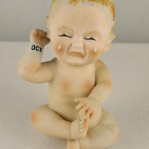 May include: A porcelain figurine of a crying baby with closed eyes and a frowning mouth. The baby is seated with one arm raised to its ear and the word "OCT" printed on its wrist. The figurine has a light skin tone with rosy cheeks and a small tuft of blonde hair.