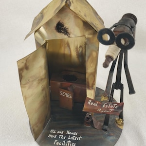 May include: A handcrafted metal sculpture of a small house with an open door, a telescope, and a sign that reads "Real Estate Agent." The house has the text "All Our Homes Have The Latest Facilities" and a "Sears" sign.