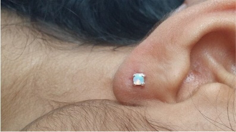 May include: A close-up of a person's ear with a small, white opal stud earring.