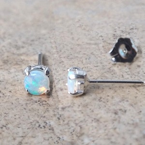 May include: A pair of opal stud earrings with a silver setting. The opals have a milky white base with flashes of blue and green. The earrings are shown with a single earring back.
