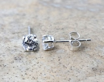 Puede incluir: Dos pendientes de botón de plata con piedras preciosas redondas y transparentes. Los pendientes tienen un diseño sencillo y clásico, con cierre de poste y mariposa. Las piedras preciosas brillan, reflejando la luz. Se muestran sobre una superficie texturizada.