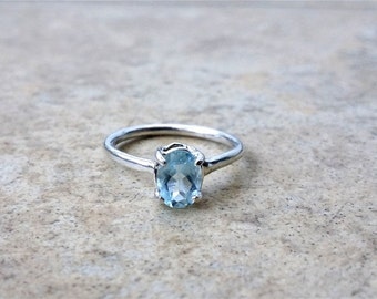 Pode incluir: Um anel de prata com uma pedra preciosa oval azul clara. A pedra é fixada em uma cravação de quatro garras, e a banda é de prata polida. O anel é exibido em uma superfície texturizada neutra.
