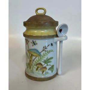 Könnte beinhalten: Ein Keramiktopf mit Deckel und kleinem Löffel. Der Topf ist weiß mit einem bemalten Pilz- und Farn-Design. Der Deckel ist braun und der Löffel hat einen roten Marienkäfer. Es ist ein dekorativer Küchenartikel.