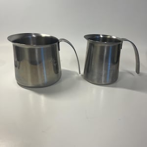 May include: Two stainless steel pitchers with curved handles. The pitchers are cylindrical with a flared rim. The metal has a brushed finish, reflecting the light. The pitchers are likely for serving beverages.