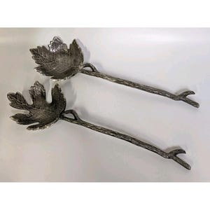 Puede incluir: Dos cucharas de servir plateadas. Cada cuchara presenta un cuenco en forma de hoja y un mango en forma de tallo con un extremo en forma de ramita. Las cucharas están diseñadas para parecerse al follaje natural.