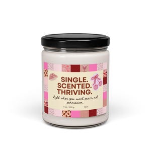 May include: A white scented candle in a glass jar with a black lid. The label features the text "SINGLE. SCENTED. THRIVING." and decorative pink and red squares. The candle is 9 oz / 255 g and has a 50-hour burn time.