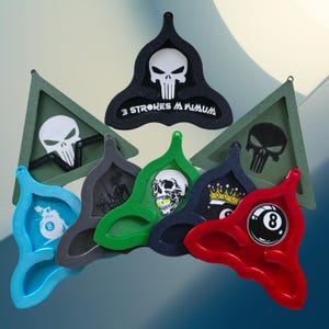 May include: A collection of colorful, decorative pool rack holders. The holders are in various shapes and colors, including blue, gray, green, navy, and red. Each holder features a unique design, such as a skull, a crown, or an eight ball.