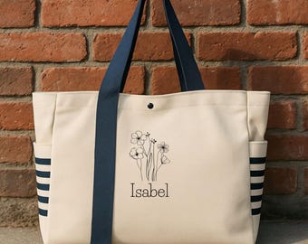 May include: Cream-colored tote bag with navy blue accents. Features a floral design and the name "Isabel" in black script. Includes navy blue striped side pockets and navy blue straps. The bag is set against a brick wall.