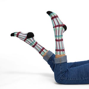 May include: A pair of crew socks featuring a white base with a plaid pattern in shades of maroon, teal, and light blue. The socks have solid black cuffs at the top and solid black toe sections. The socks are worn with blue denim jeans.