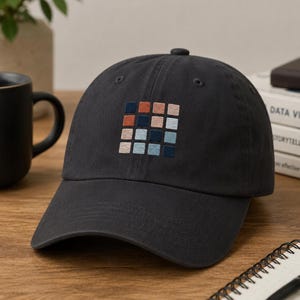 May include: A dark gray baseball cap with a square embroidered design of various colored squares. The squares are in shades of orange, blue, and light pink. The cap has a curved brim and a button on top.