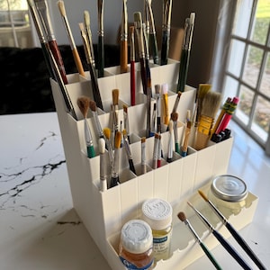 May include: A cream-colored, tiered art supply organizer filled with various paint brushes of different sizes and shapes. Two small bottles of paint and a jar of medium are also visible. The brushes are arranged neatly, ready for use.