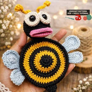 Puede incluir: Un juguete de abeja de ganchillo hecho a mano con un cuerpo negro, vientre a rayas amarillas y negras y alas azul claro. La abeja tiene ojos blancos grandes con pupilas negras, boca rosa y antenas amarillas. El texto "CROCHET PATTERN: PDF + VIDEO TUTORIAL" es visible.