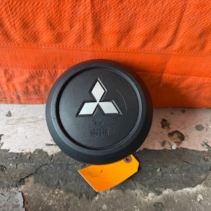 Puede incluir: Emblema negro del volante Mitsubishi con logotipo plateado. El emblema es redondo con una superficie texturizada y el texto "SRS AIRBAG". Una etiqueta naranja está adherida al emblema.