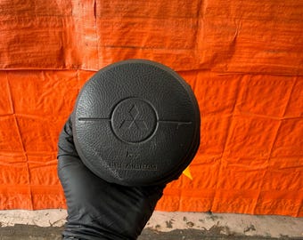Può includere: Coperchio airbag del volante nero con il logo Mitsubishi. Il coperchio è rotondo e ha una superficie strutturata. Le parole "AIRBAG" sono stampate nella parte inferiore del coperchio.