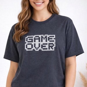 Op de afbeelding: Donkergrijs t-shirt met de pixelachtige witte tekst "GAME OVER". Het shirt met korte mouwen is gemaakt van een zacht materiaal en is geschikt voor casual wear. Het ontwerp heeft een retro videogame-esthetiek.