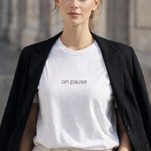 Op de afbeelding: Een wit t-shirt met de woorden "on pause" in het zwart gedrukt, gedragen met een zwarte blazer en een lichtgekleurde broek. Het model draagt kleine oorringen en een delicate ketting.