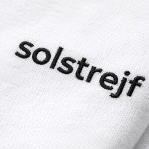 Close-up of black embroidery “solstrejf” on white cotton t-shirt, detailed stitch texture, minimalist text design