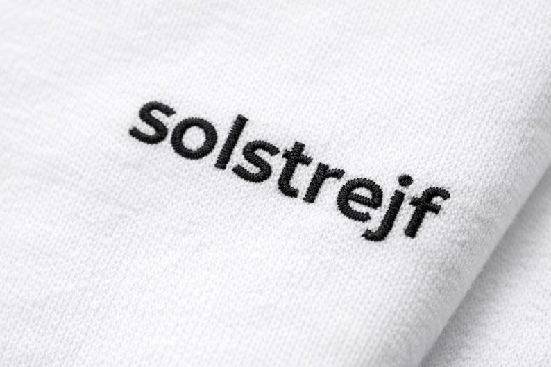 Close-up of black embroidery “solstrejf” on white cotton t-shirt, detailed stitch texture, minimalist text design