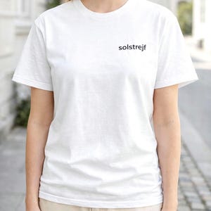 May include: A white crew neck t-shirt with the word "solstrejf" printed in black on the chest. The shirt is worn by a person, paired with beige shorts. The background is a blurred outdoor scene.