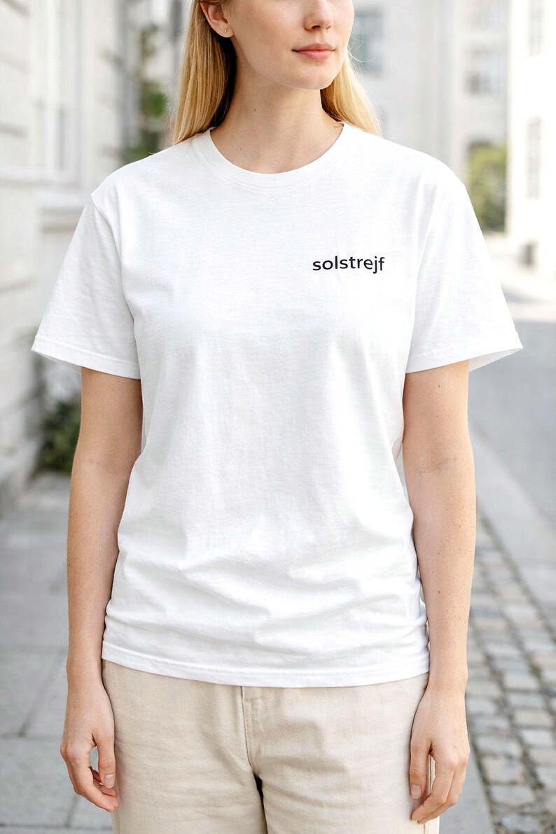 May include: A white crew neck t-shirt with the word "solstrejf" printed in black on the chest. The shirt is worn by a person, paired with beige shorts. The background is a blurred outdoor scene.