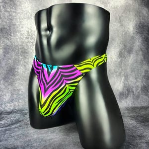 Puede incluir: Unos calzoncillos tipo bikini con un vibrante estampado de rayas de cebra en verde neón, rosa brillante y azul. La prenda presenta una cintura de tiro bajo y un diseño de pierna alta, destacando un llamativo estampado animal multicolor.