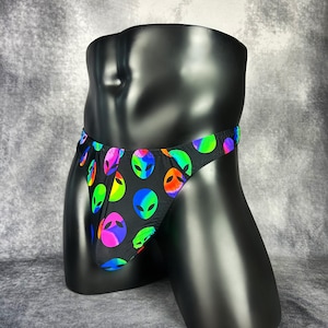 May include: A black thong featuring a repeating pattern of alien heads in vibrant, multi-coloured gradients of neon green, pink, blue, and orange. The garment has a low-rise waistband and a contoured front pouch design with visible stitching.