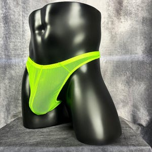Puede incluir: Tanga de color verde neón sobre un maniquí negro. La ropa interior está hecha de una tela transparente y ligera con una cinturilla a juego. El maniquí se encuentra sobre un fondo gris texturizado.