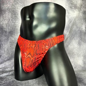 May include: A pair of underwear featuring a vibrant red and black snakeskin pattern. The garment has a low-rise waistband and a form-fitting design that covers the front, displayed on a smooth, black, headless torso mannequin form.