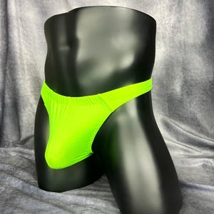 May include: Neon green thong underwear displayed on a black mannequin. The underwear has a thin waistband and a minimal back coverage. The fabric appears to be a stretchy material.