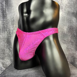 Può includere: Un costume da bagno slip rosa acceso dall'aspetto metallico, esposto su un manichino nero. Il costume da bagno ha una vita bassa e una vestibilità aderente. Il tessuto presenta un motivo a pois strutturato. Il manichino è posizionato su uno sfondo grigio.