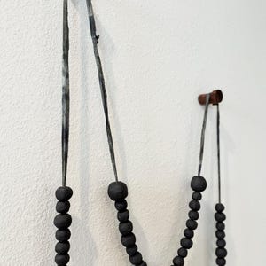 K&ouml;nnte beinhalten: Drei Str&auml;nge schwarzer Perlendekoration, die an Holzd&uuml;beln an einer wei&szlig;en Wand h&auml;ngen. Die Perlen variieren in der Gr&ouml;&szlig;e und sind auf dunklen Schn&uuml;ren aufgef&auml;delt. Die Holzd&uuml;bel sind zylindrisch und scheinen aus dunklem Holz zu sein.