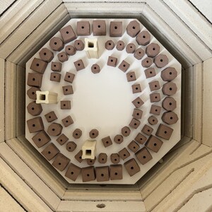 K&ouml;nnte beinhalten: Innenansicht eines Brennofens mit Keramikst&uuml;tzen. Die St&uuml;tzen haben verschiedene Formen und Gr&ouml;&szlig;en, darunter W&uuml;rfel, Zylinder und Achtecke. Die St&uuml;tzen sind auf einem wei&szlig;en Keramikregal angeordnet. Der Innenraum des Brennofens besteht aus beigen Ziegeln.