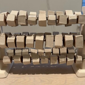 K&ouml;nnte beinhalten: Eine Keramik-Abakus-Skulptur mit drei Reihen rechteckiger Bl&ouml;cke in verschiedenen Beige- und Graut&ouml;nen. Die Bl&ouml;cke sind auf d&uuml;nnen Metallst&auml;ben aufgereiht und werden von einem wei&szlig;en Sockel getragen. Die Skulptur ist eine moderne Interpretation eines klassischen Lernwerkzeugs.