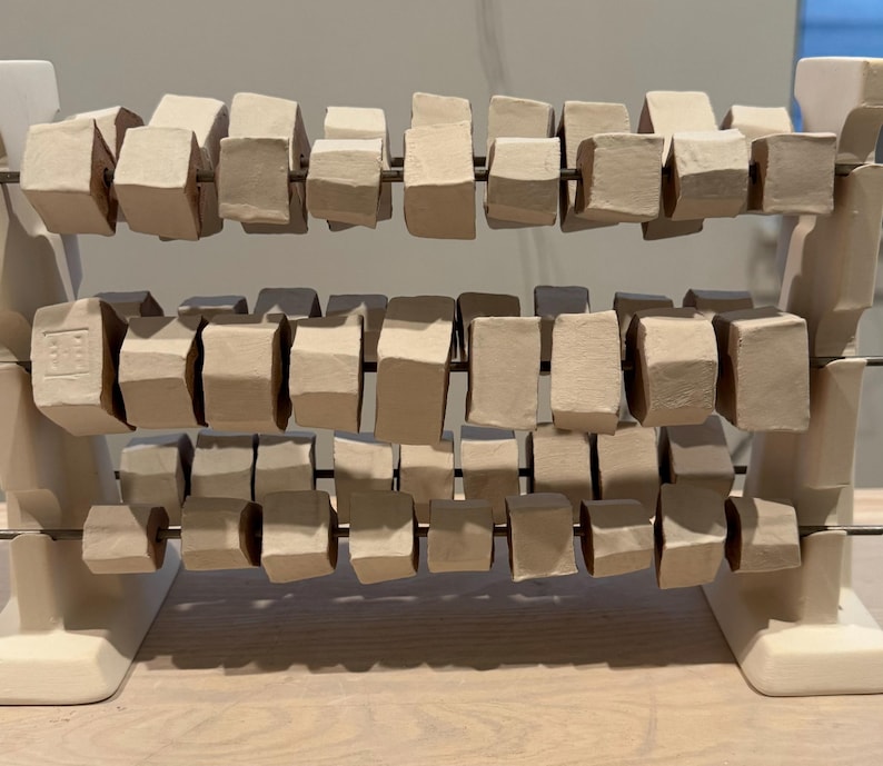 K&ouml;nnte beinhalten: Eine Keramik-Abakus-Skulptur mit drei Reihen rechteckiger Bl&ouml;cke in verschiedenen Beige- und Graut&ouml;nen. Die Bl&ouml;cke sind auf d&uuml;nnen Metallst&auml;ben aufgereiht und werden von einem wei&szlig;en Sockel getragen. Die Skulptur ist eine moderne Interpretation eines klassischen Lernwerkzeugs.