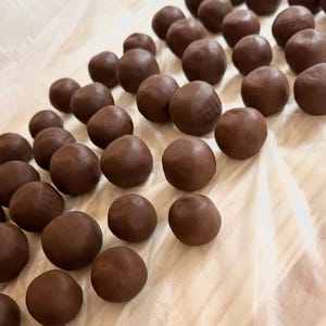 K&ouml;nnte beinhalten: Nahaufnahme von zahlreichen dunkelbraunen, kugelf&ouml;rmigen Objekten, die Tr&uuml;ffeln oder Pralinen &auml;hneln, auf einer wei&szlig;en Oberfl&auml;che. Einige Kugeln weisen strukturierte Muster auf. Die Beleuchtung betont die glatte Textur der Leckereien.